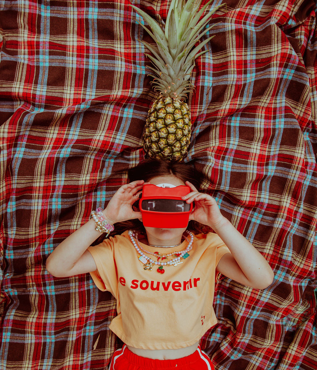 girl with red camera and pineapple wearing charm necklace and bracelet charms by roger. cherry charms, bow charm, gummy bear charm, chilli charm, ice cream charm. shop charms and jewellery.