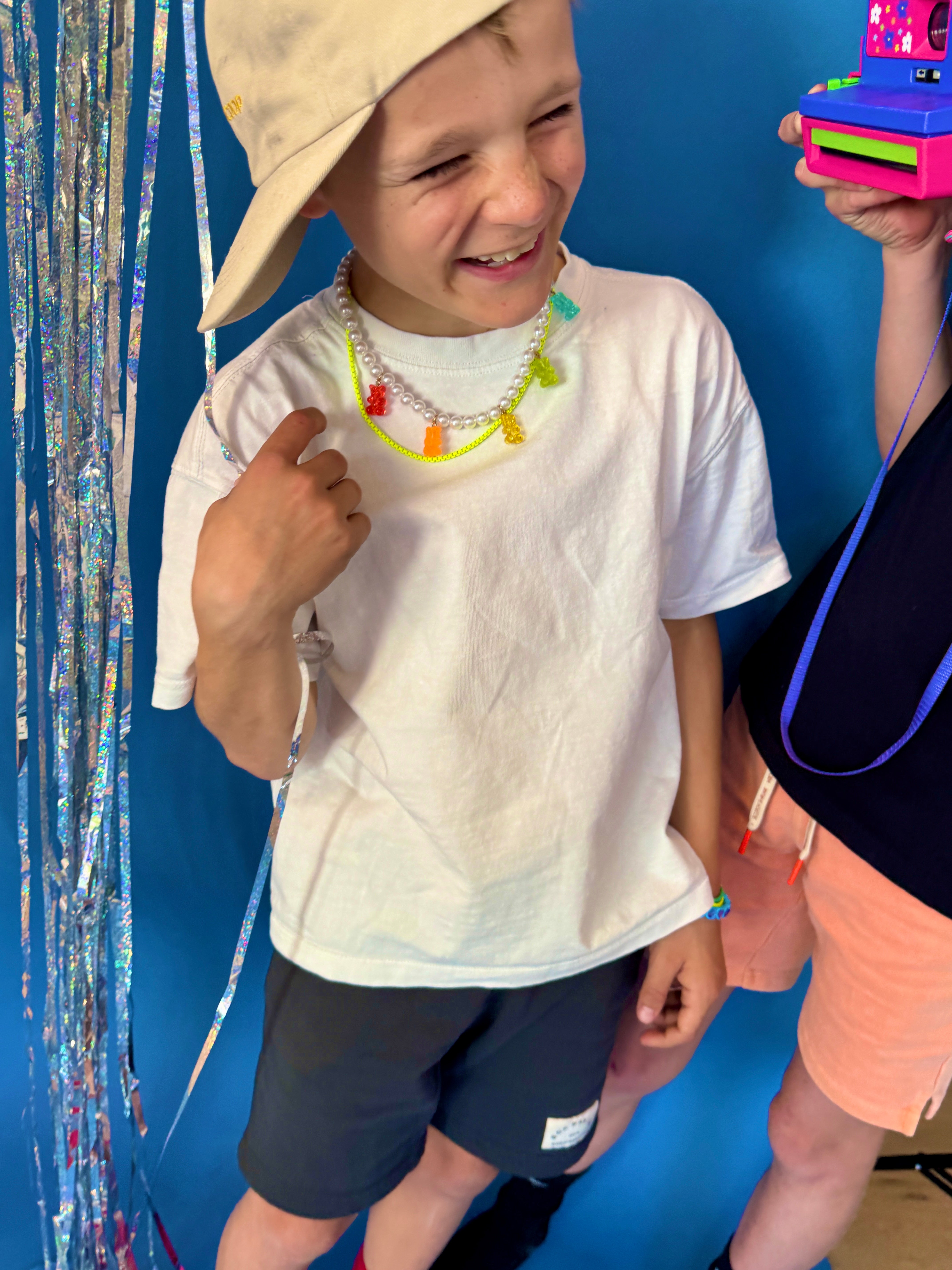 Child wearing a beige hat and white shirt with colorful accessories, standing next to a person holding a camera against a blue background.