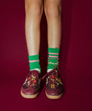 A pair of feet wearing green socks with shoe charms attached to the laces, along with red adidas sambas. Sneaker charms include gold bow charm, heart charm, key charm and pearl charm.