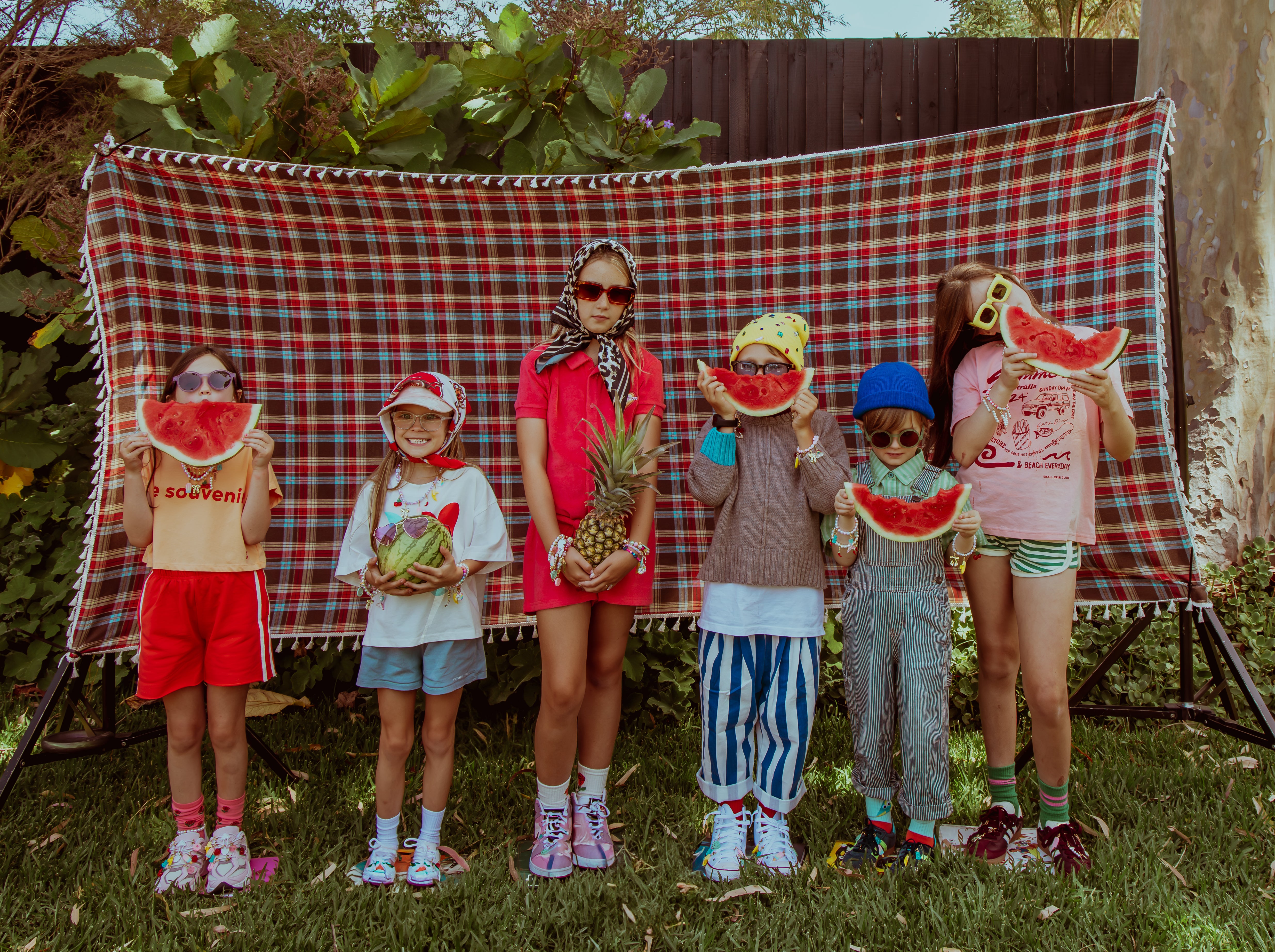 Children posing with watermelon-themed props in front of a plaid blanket outdoors.