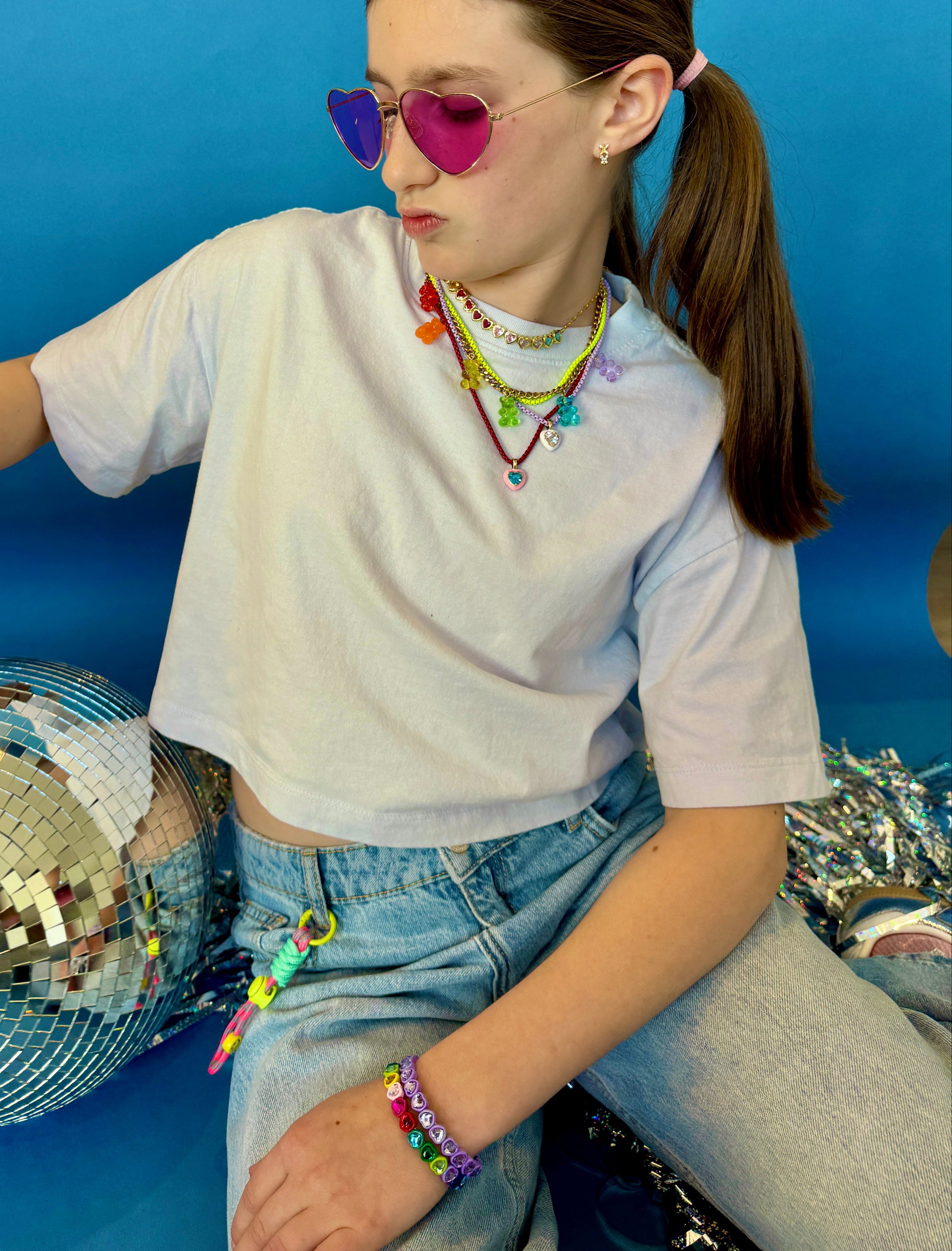 girl wearing colourful sunglasses and multiple necklaces, sitting with a disco ball against a blue background