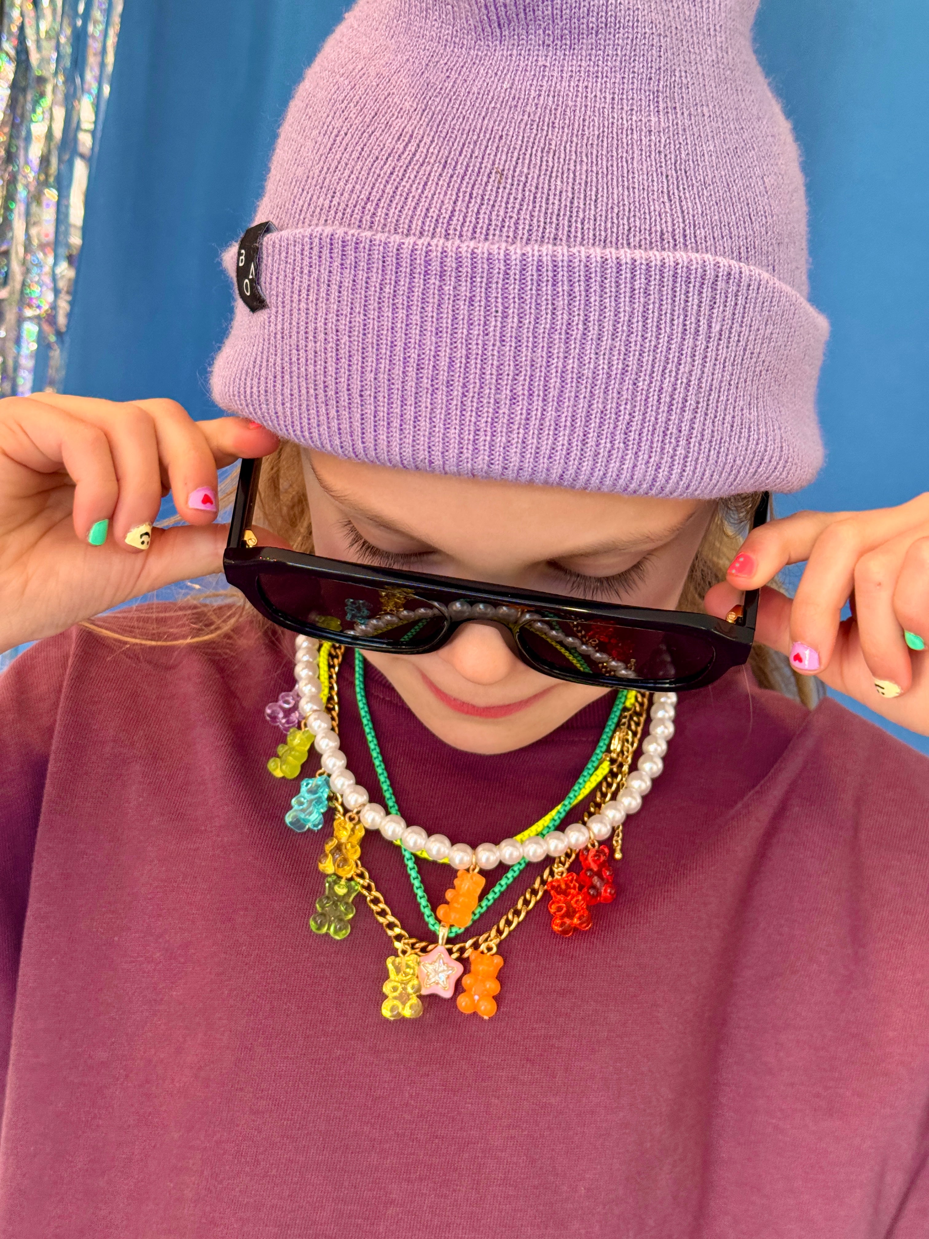 Person wearing a purple beanie and colorful necklace with sunglasses, against a blue background