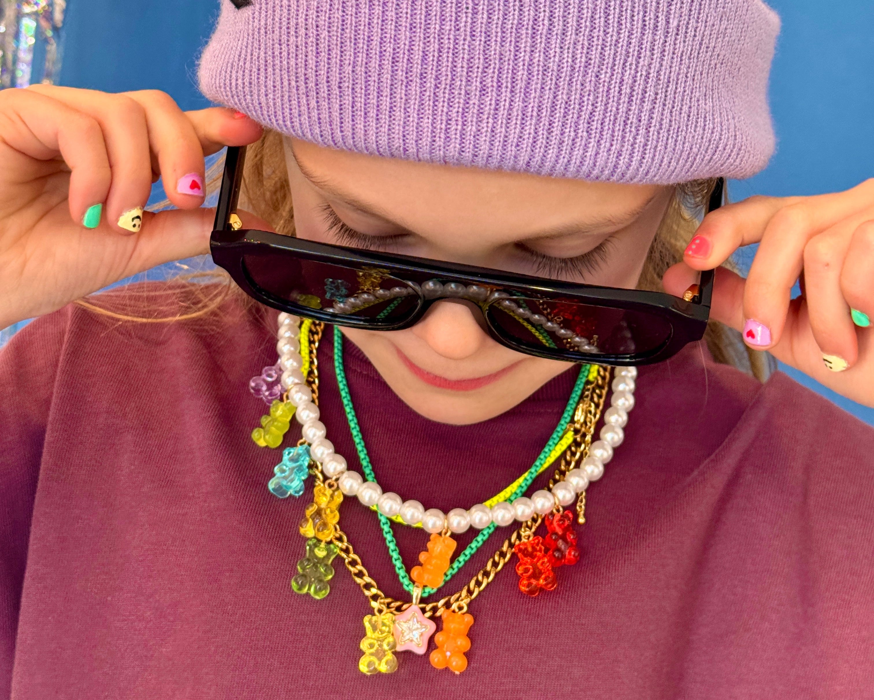 Person wearing a purple beanie and colorful necklace with sunglasses, against a blue background